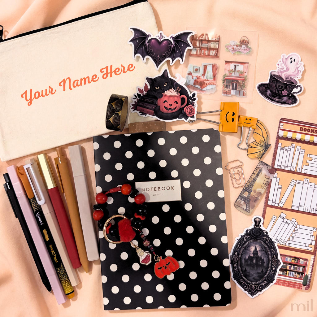 A styled flat-lay of stationery and desk accessories on a peach fabric background, including a cream zip pouch with orange text, a black-and-white polka dot notebook, pens, stickers, bookmarks, and small decorative items.