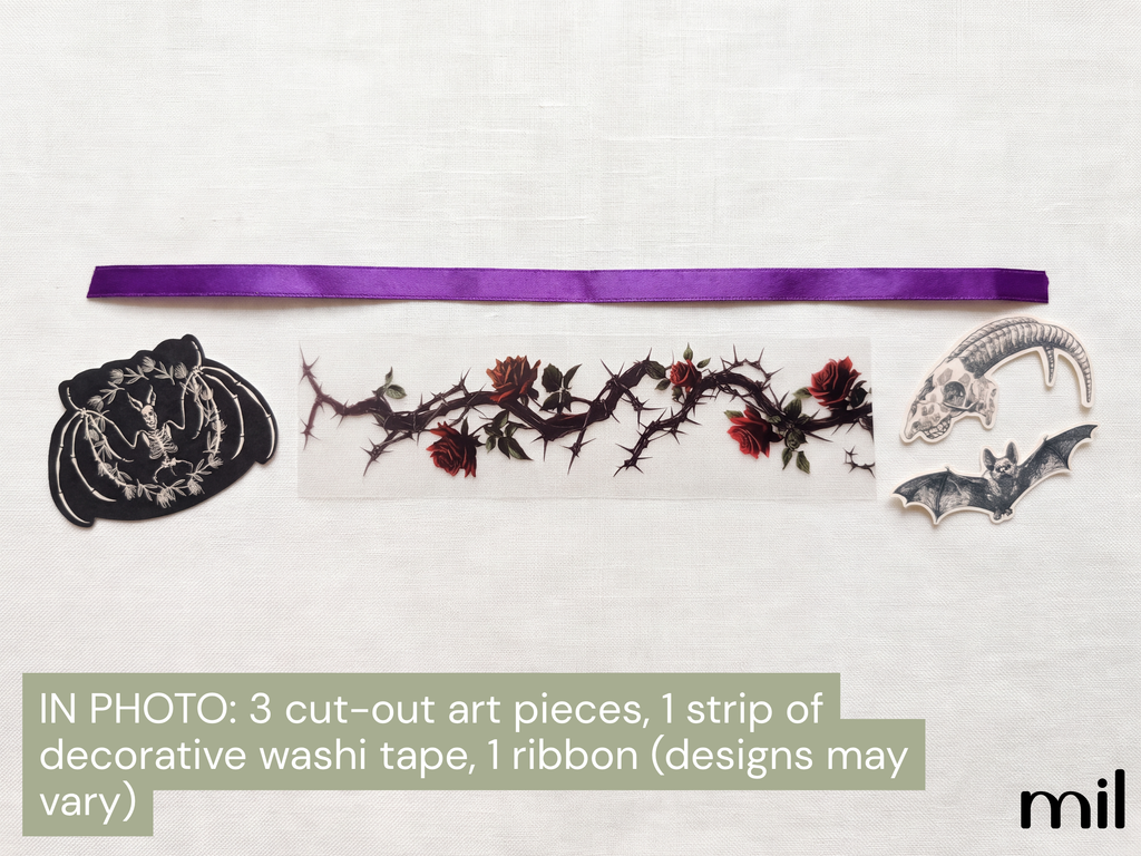 A flat lay of craft supplies on a white textured background, including a purple ribbon, a strip of decorative washi tape with a thorny rose design, a black embroidered patch, and two gray bat-shaped cutouts. Text at the bottom reads 'IN PHOTO: 3 cut-out art pieces, 1 strip of decorative washi tape, 1 ribbon (designs may vary)'.