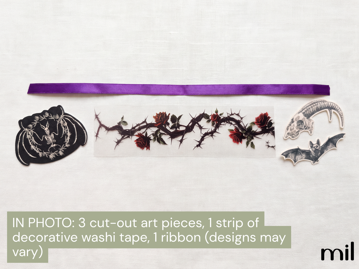 A flat lay of craft supplies on a white textured background, including a purple ribbon, a strip of decorative washi tape with a thorny rose design, a black embroidered patch, and two gray bat-shaped cutouts. Text at the bottom reads 'IN PHOTO: 3 cut-out art pieces, 1 strip of decorative washi tape, 1 ribbon (designs may vary)'.