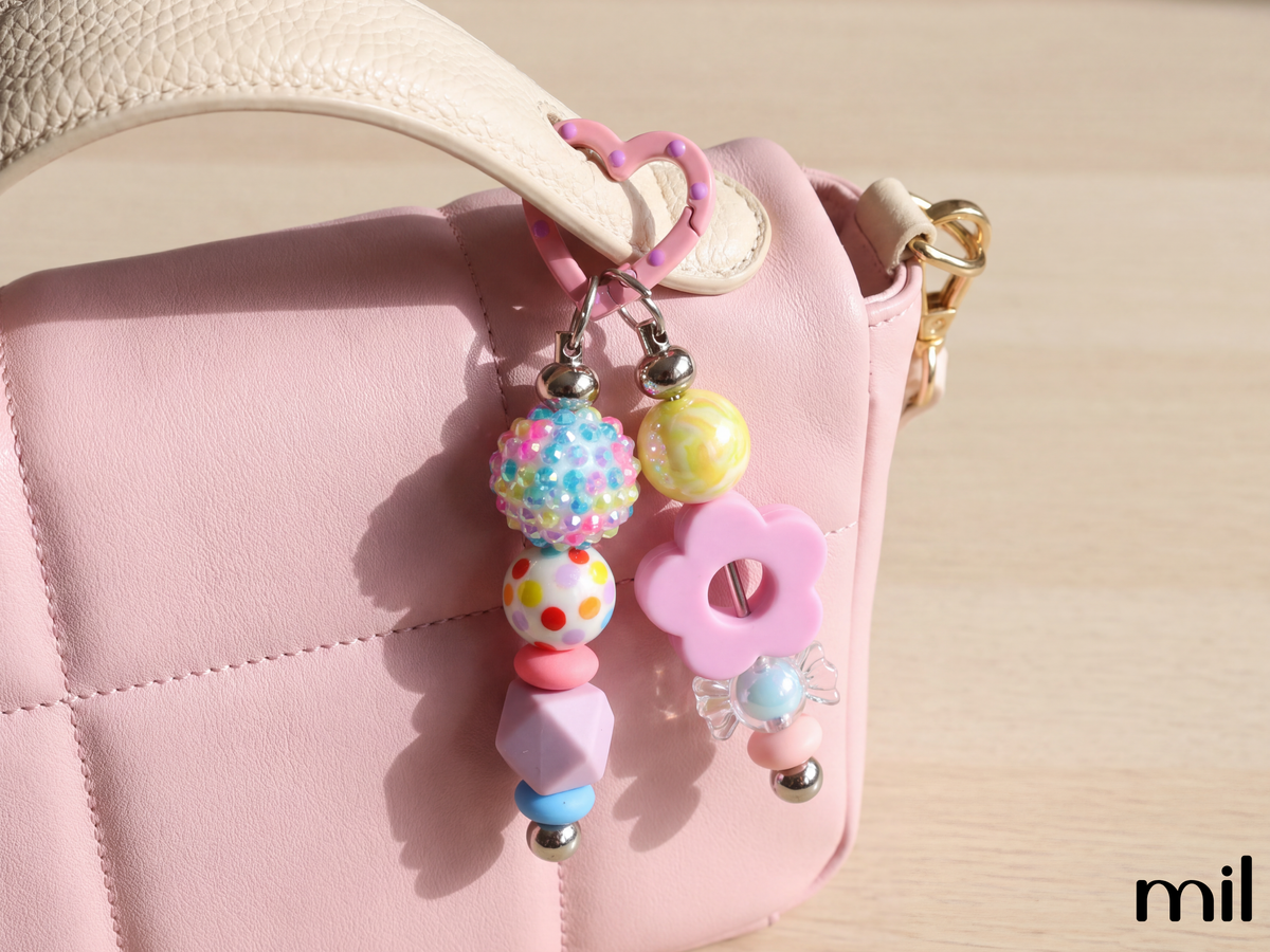 A pastel pink quilted handbag with a cream handle, decorated with a colorful charm keychain featuring pastel beads, a pink flower charm, and a pink heart ring, shown on a light wood background.