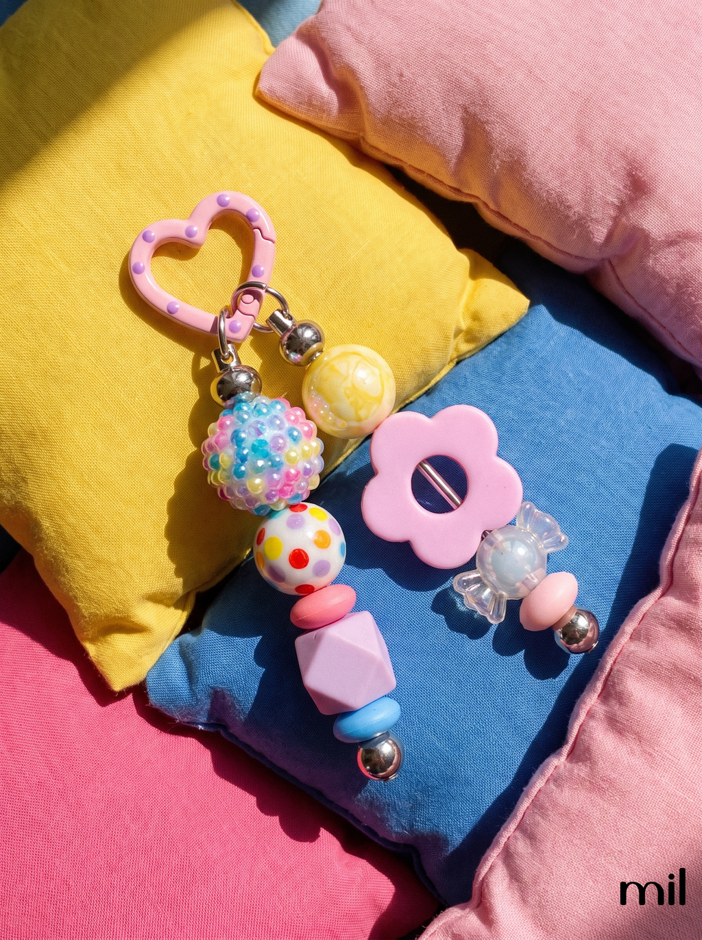 A pastel keychain set styled on colorful pillows. The set includes a pink heart-shaped key ring, a beaded keychain with a glittery multicolor ball, a yellow ball, and pink and blue beads, and a pink flower-shaped teether with a clear bead.