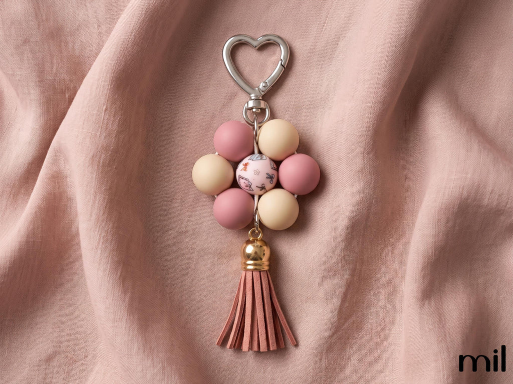 A floral bead keychain with a silver heart-shaped clasp, featuring pink and cream beads, a marbled center bead, and a pink leather tassel, displayed on draped dusty pink fabric.