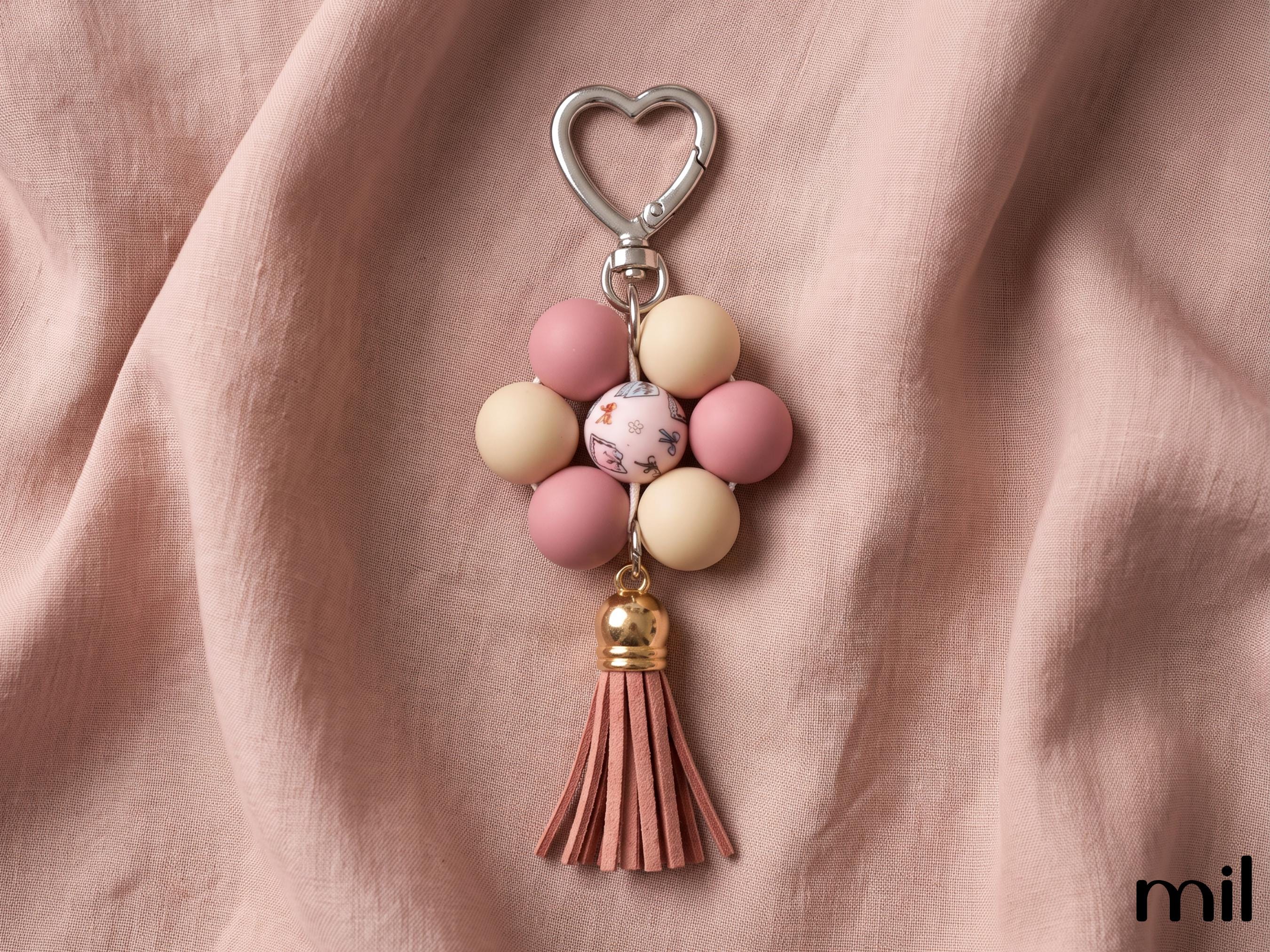 A floral bead keychain with a silver heart-shaped clasp, featuring pink and cream beads, a marbled center bead, and a pink leather tassel, displayed on draped dusty pink fabric.