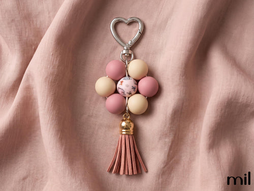 A floral bead keychain with a silver heart-shaped clasp, featuring pink and cream beads, a marbled center bead, and a pink leather tassel, displayed on draped dusty pink fabric.