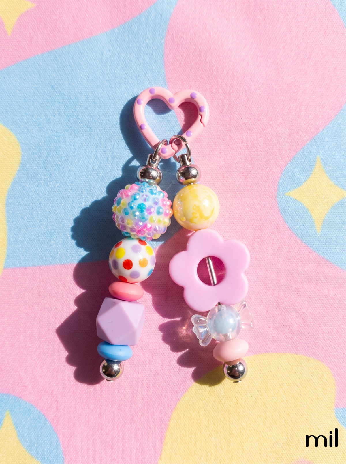 A pastel pink and multicolor beaded keychain charm with a heart-shaped ring, flower bead, and colorful patterned beads, photographed on a pastel pink, blue, and yellow patterned background.