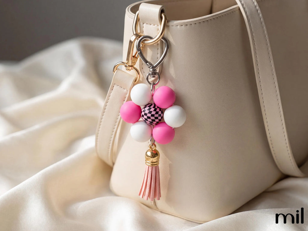 A close-up of a cream-colored leather handbag with a decorative keychain charm hanging from the ring. The charm features pink and white beads, a small black-and-white checkered center, and a pink tassel, set against draped cream fabric.