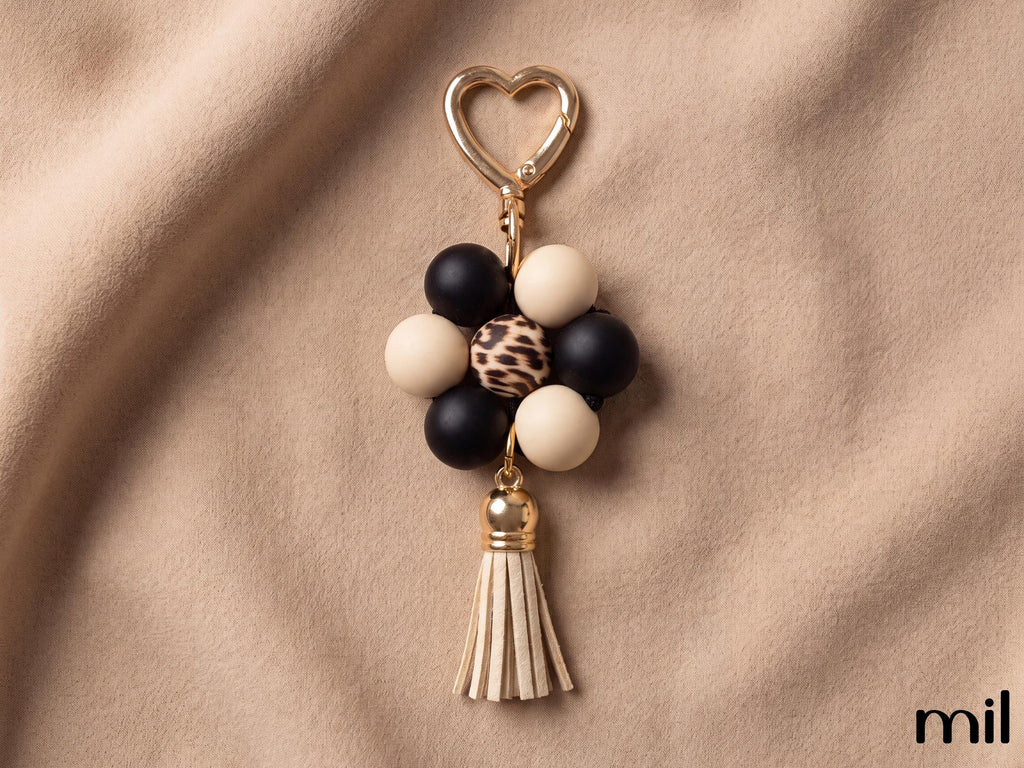 A beaded flower-shaped keychain charm with black and cream beads, a leopard-print center bead, a gold heart clasp, and a cream tassel, displayed on a draped beige fabric background.
