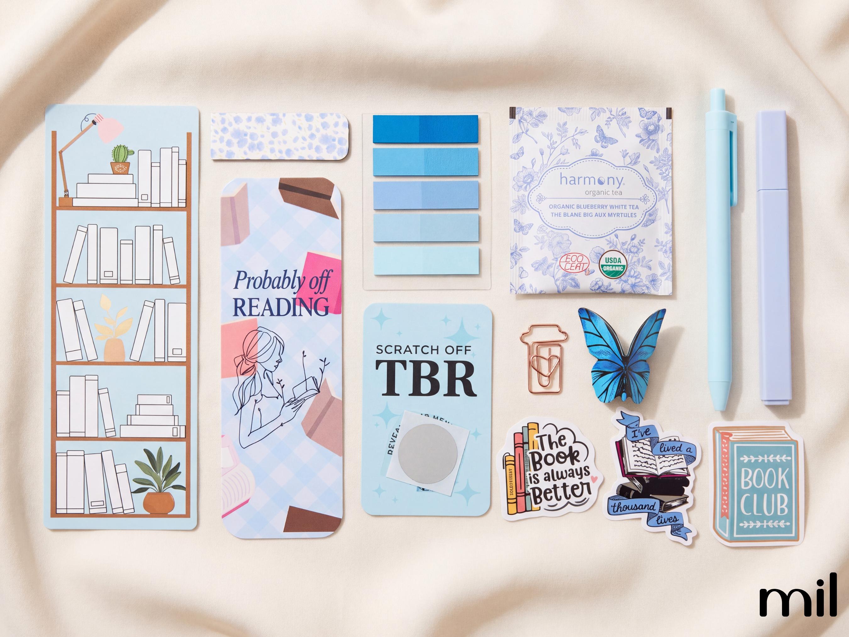 A flat-lay collection of pastel blue and lavender stationery and sticker accessories on a cream fabric background, including a bookshelf bookmark, sticky notes, a notepad, a pen, a highlighter, and several decorative stickers with book-themed illustrations.