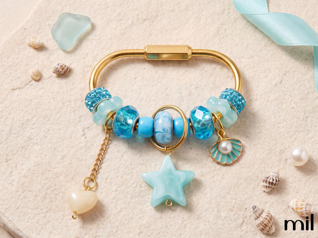 A gold-tone charm bracelet styled with blue and turquoise beads, a star-shaped charm, a shell charm, and a dangling pearl bead, arranged on a textured beige surface with seashells and a light blue ribbon.