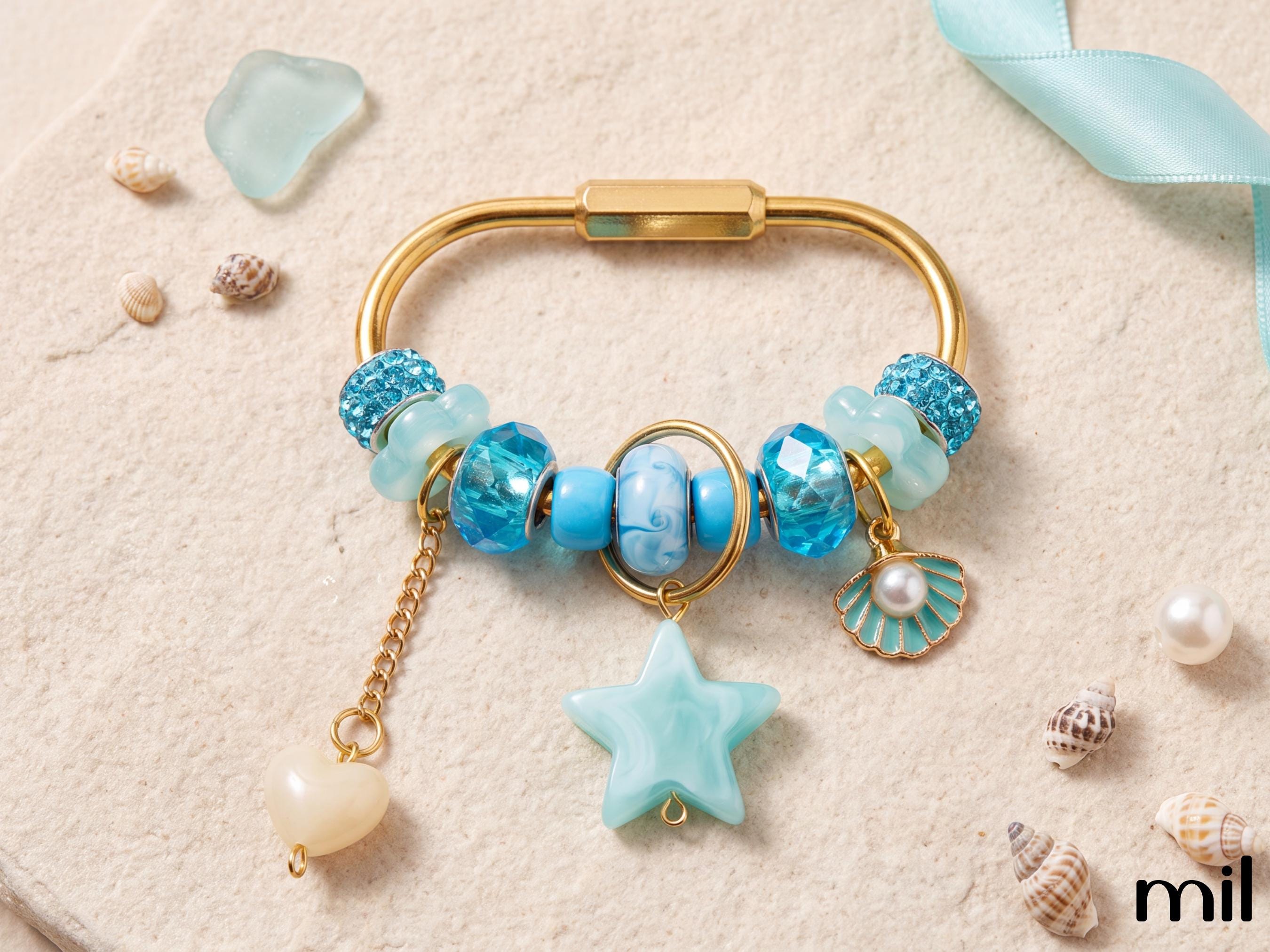 A gold-tone charm bracelet styled with blue and turquoise beads, a star-shaped charm, a shell charm, and a dangling pearl bead, arranged on a textured beige surface with seashells and a light blue ribbon.