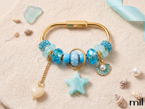 A gold-tone charm bracelet styled with blue and turquoise beads, a star-shaped charm, a shell charm, and a dangling pearl bead, arranged on a textured beige surface with seashells and a light blue ribbon.