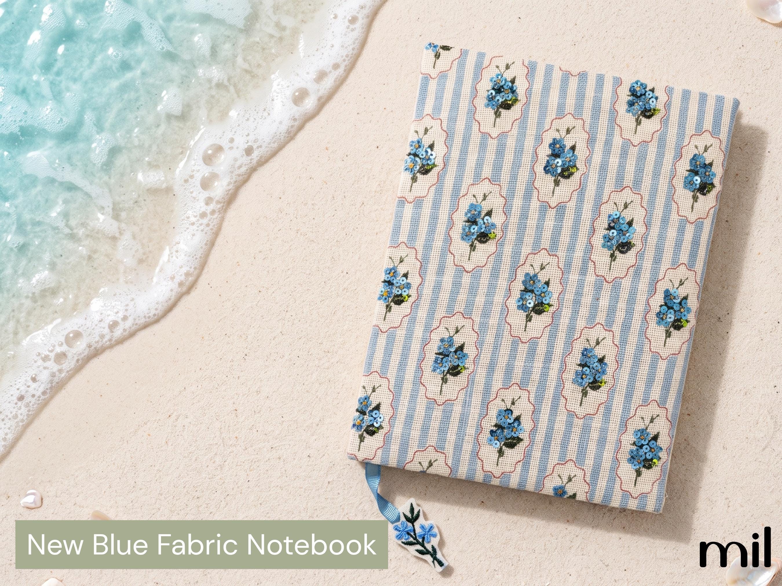 A fabric notebook with a blue floral and light blue striped pattern lies on a sandy beach next to the ocean water. A green banner at the bottom left reads 'New Blue Fabric Notebook' and a 'mil' logo is visible in the bottom right corner.