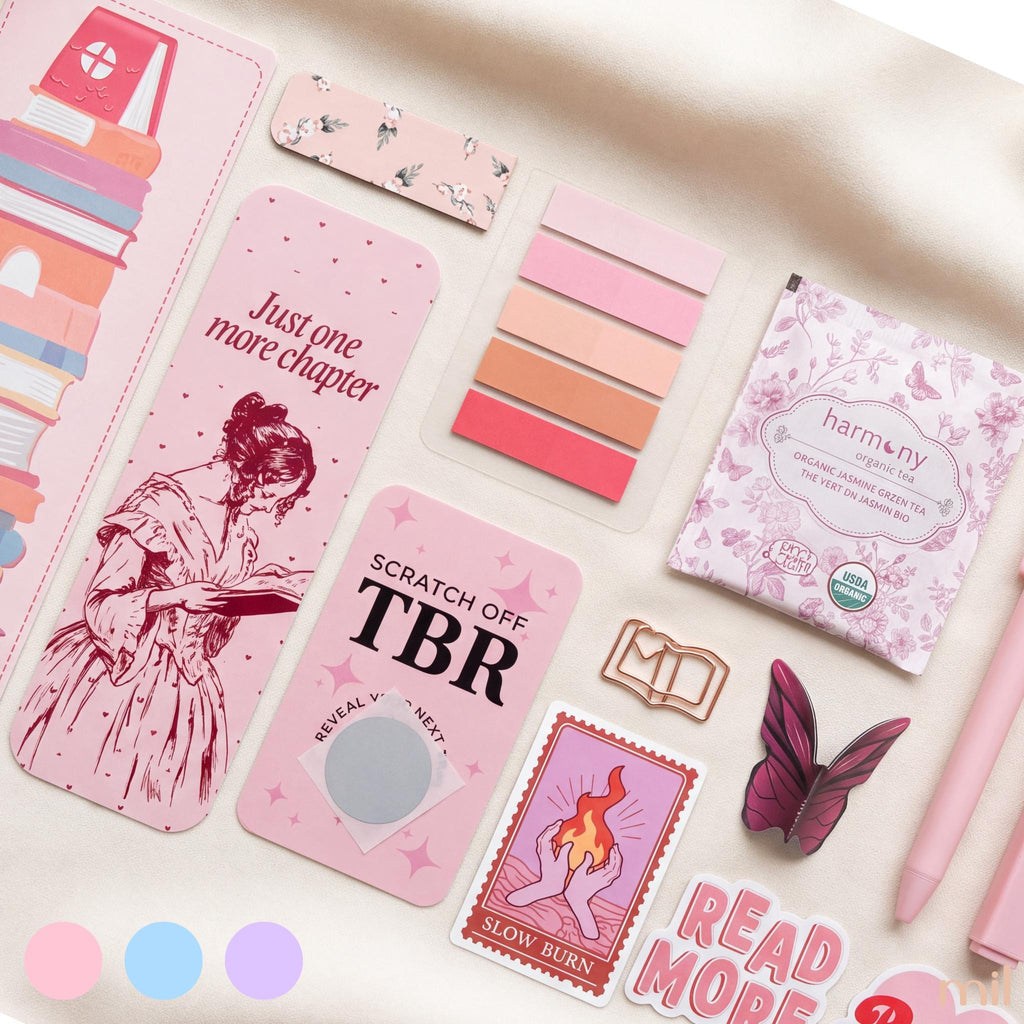 A styled flat-lay of pink stationery and desk accessories on a light fabric background, including a long bookmark with a book illustration, sticky notes, a TBR card, a notebook, a pen, and decorative paper items.