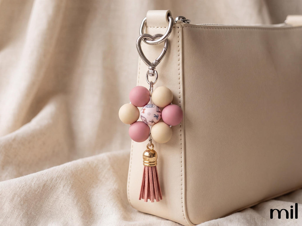 A close-up of a beige leather handbag with a decorative keychain charm featuring pink and cream beads, a floral center, and a pink tassel, set against a soft beige fabric background.