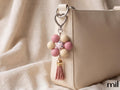 A close-up of a beige leather handbag with a decorative keychain charm featuring pink and cream beads, a floral center, and a pink tassel, set against a soft beige fabric background.