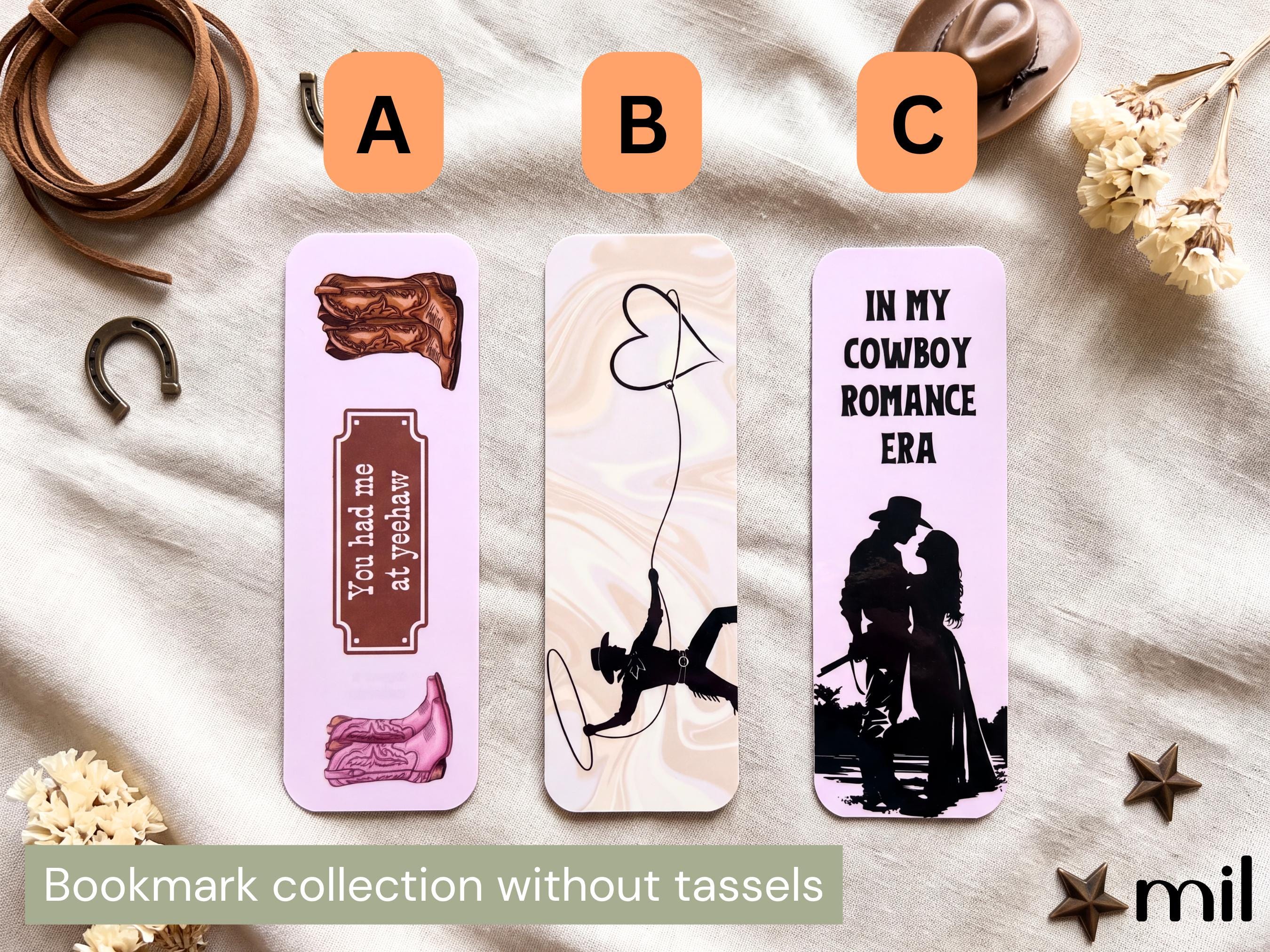 Three cowboy-themed bookmarks labeled A, B, and C displayed on a cream fabric background with western props. The bookmarks feature illustrations and text including a boot, a heart-shaped string, and a cowboy silhouette with the phrase 'IN MY COWBOY ROMANCE ERA'.