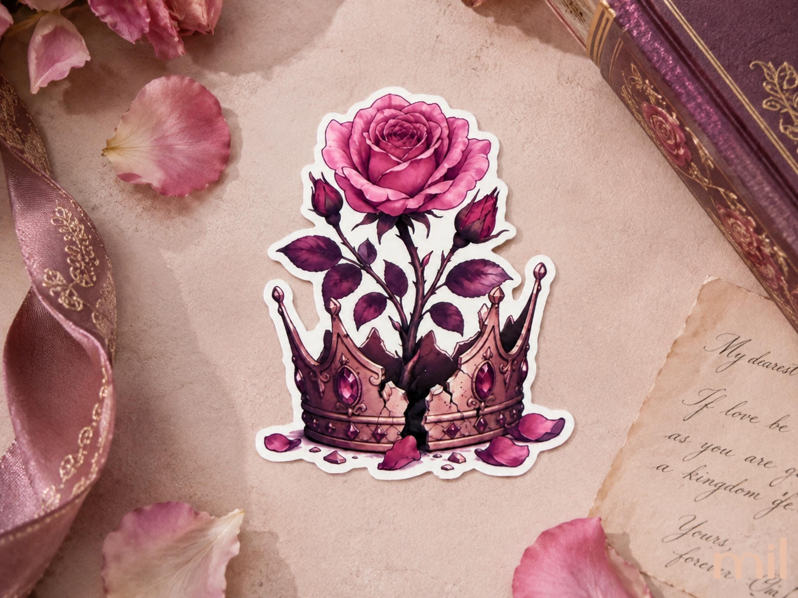A die-cut sticker featuring a pink rose blooming from a jeweled crown, displayed on a styled flat-lay background with pink rose petals, a pink ribbon, and an antique book.