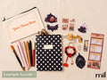 A flat-lay display of a customizable stationery and desk accessory bundle on a beige fabric background. The bundle includes a cream canvas pouch with orange text, a black and white polka dot notebook, paintbrushes, pens, stickers, a beaded keychain, and book-themed stickers.