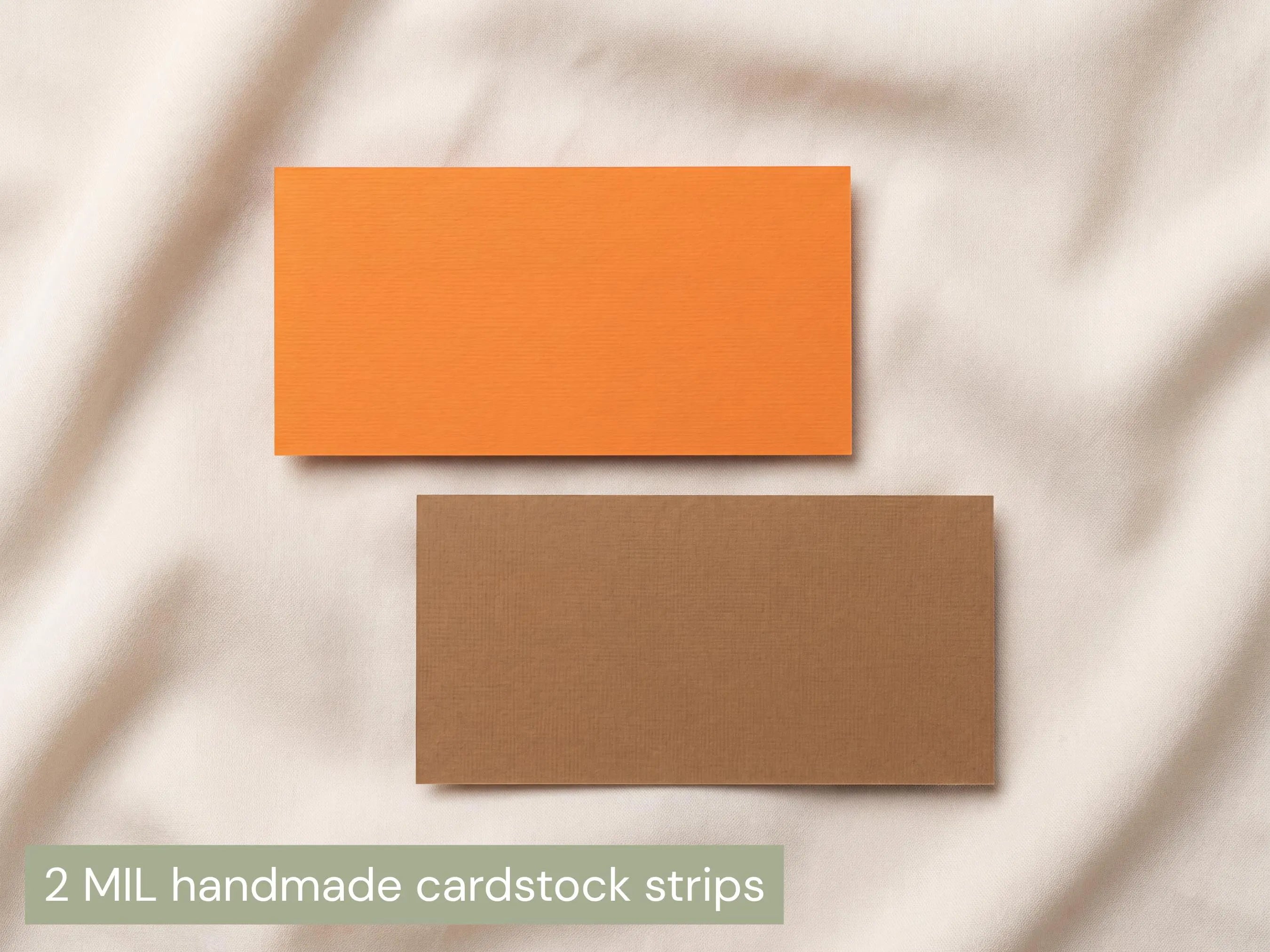 Two rectangular cardstock strips, one orange and one brown, laid flat on a soft beige fabric background. Text at the bottom reads '2 MIL handmade cardstock strips'.