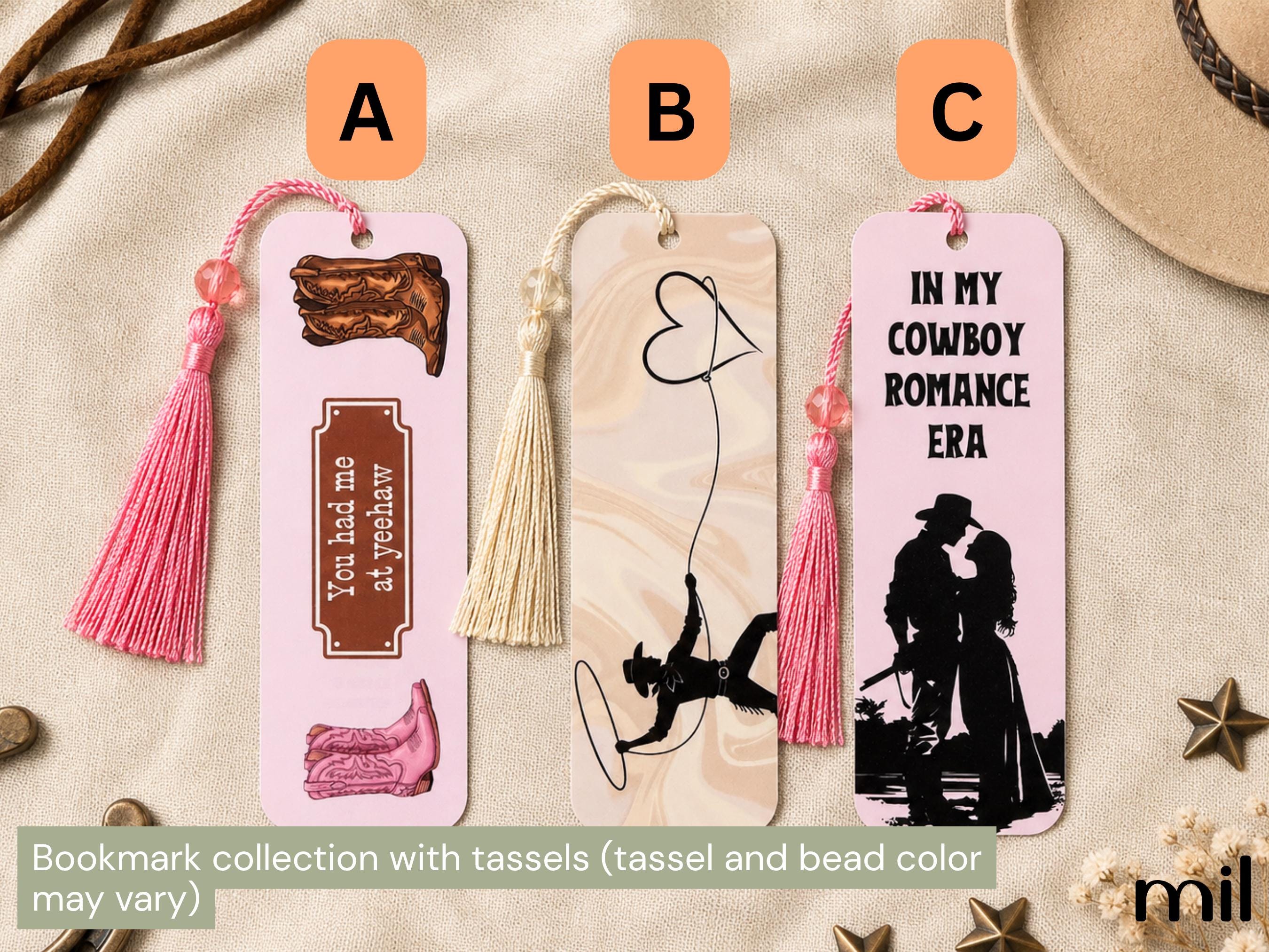 Three decorative bookmark designs labeled A, B, and C on a beige fabric background with western-themed props. The bookmarks have pink tassels and printed cow romance-themed graphics, including boots, a heart-shaped balloon, and a cowboy-and-lover silhouette with the text 'IN MY COWBOY ROMANCE ERA.
