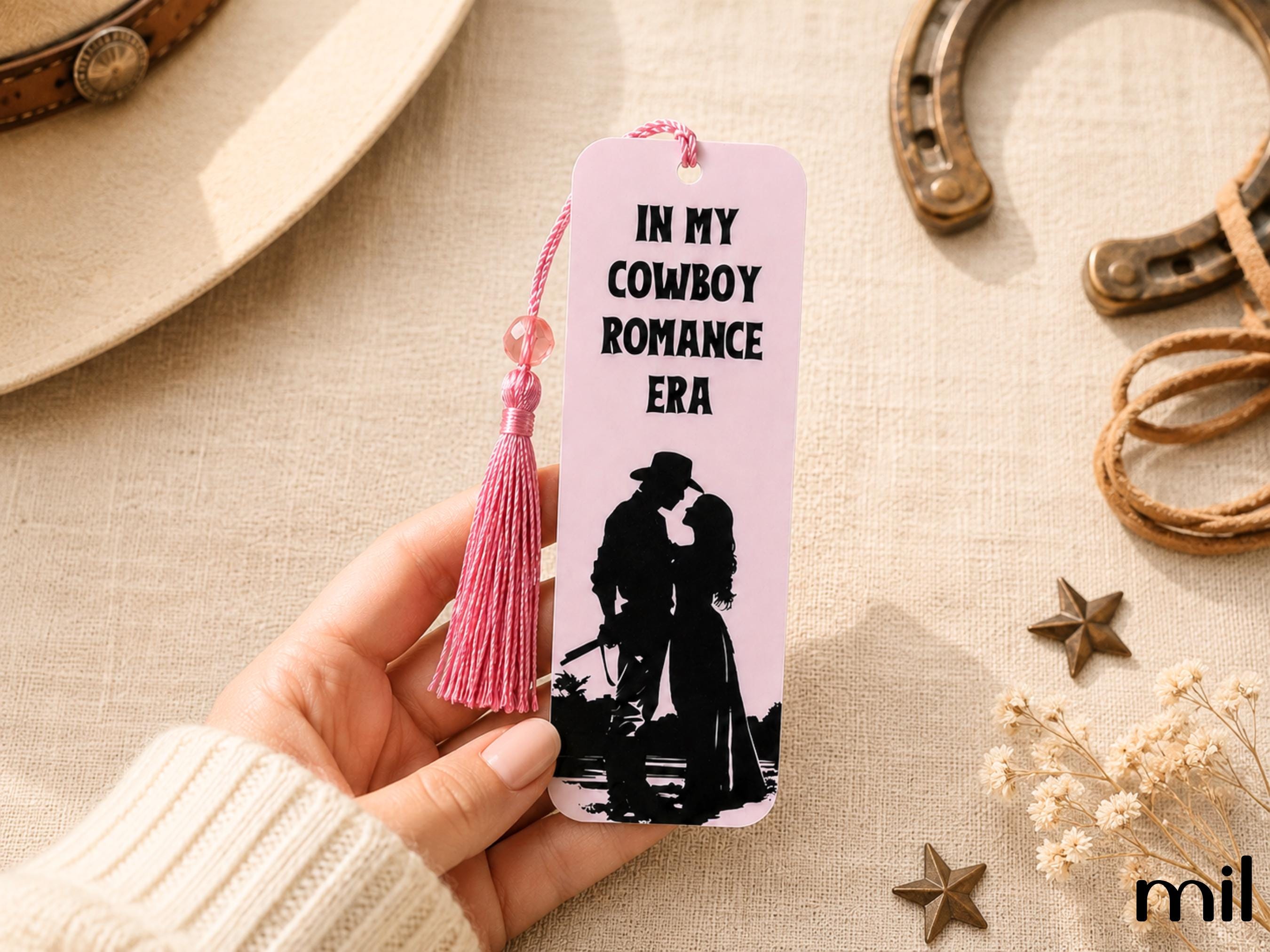 A hand holds a pink bookmark with a tassel and the text 'IN MY COWBOY ROMANCE ERA' above a black silhouette of a cowboy and cowboy-style dress figure. The scene is styled with a hat, horseshoes, rope, dried flowers, and star decorations on a beige fabric background.