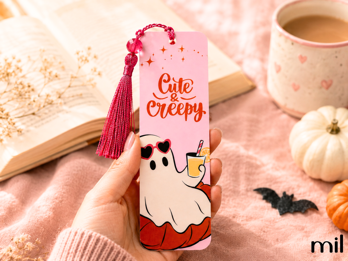 A hand holds a pink Halloween-themed metal bookmark with a tassel. The bookmark features a cute cartoon ghost wearing red sunglasses and a red pumpkin body, with the text 'Cute & Creepy' in red.