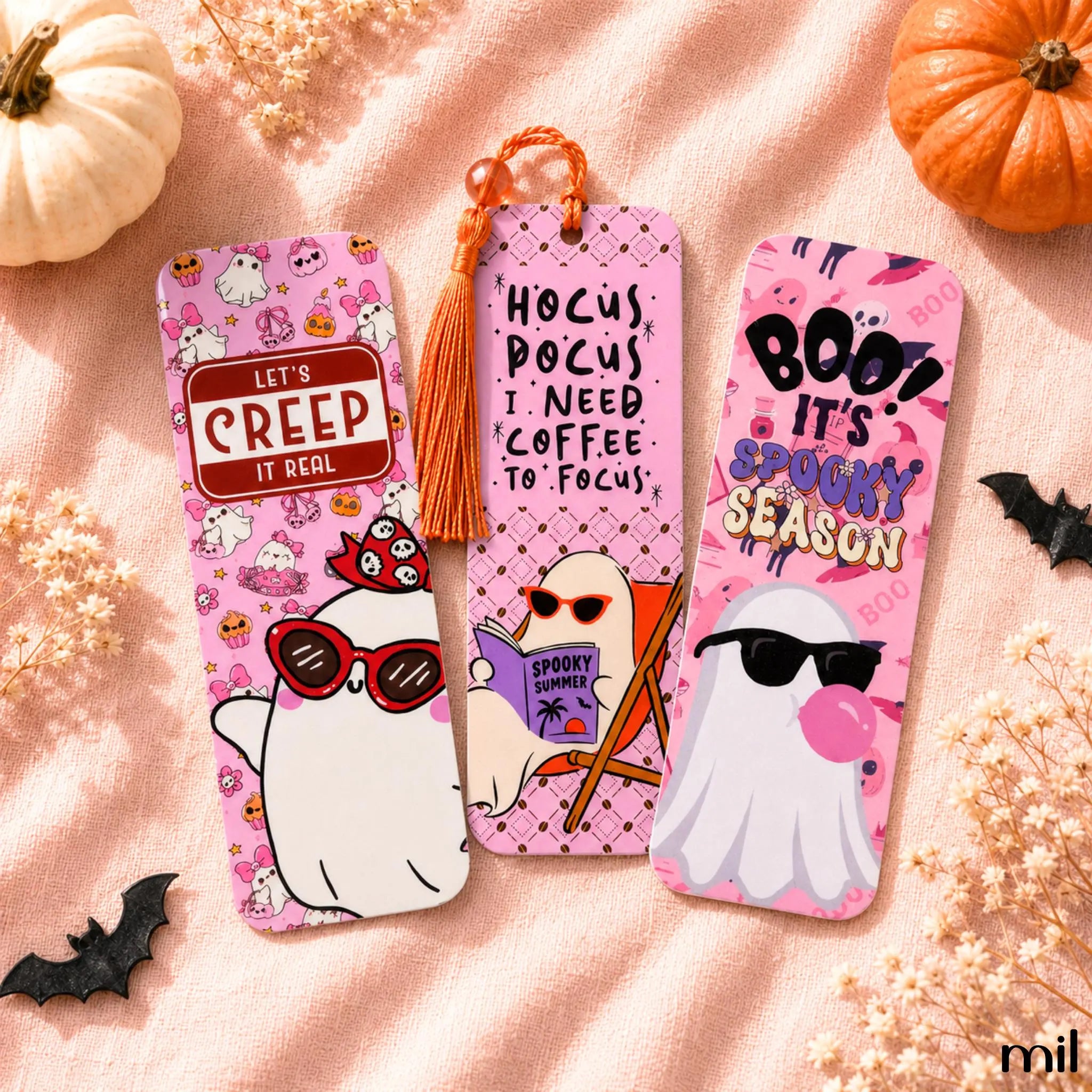 Three pink Halloween-themed paper bookmarks with cartoon ghost illustrations and decorative text, arranged on a peach fabric background with pumpkins, dried flowers, and black bat cutouts.