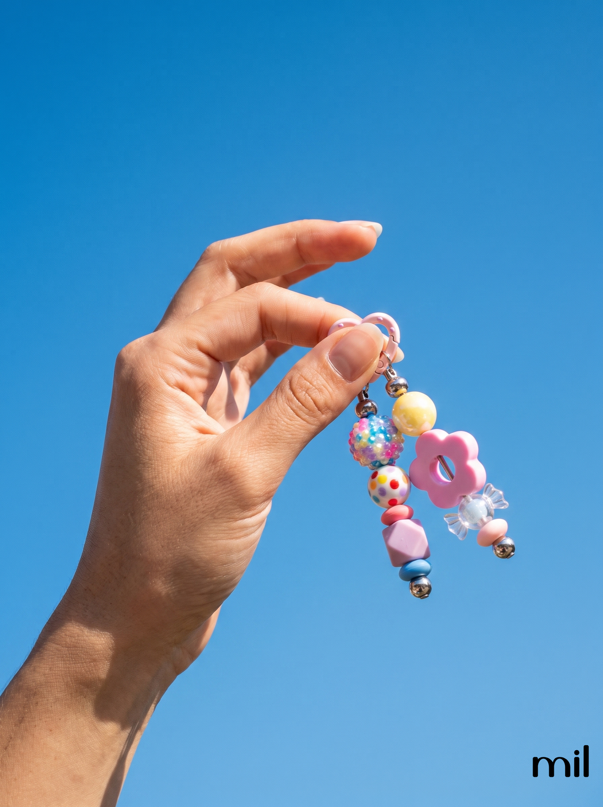 A hand holding a pastel-colored beaded keychain charm against a clear blue sky. The charm features a pink flower, a yellow bead, a pink heart, and colorful patterned beads 