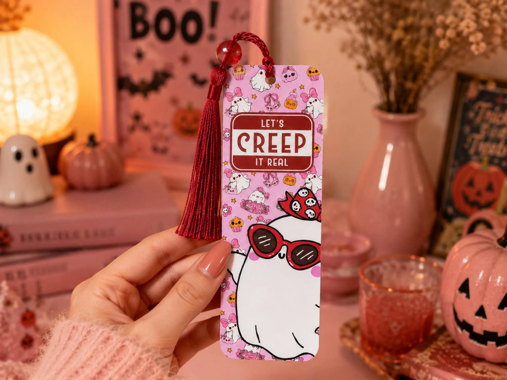 A hand holds a pink Halloween-themed bookmark featuring a cartoon ghost wearing red sunglasses and the text 'LET'S CREEP IT REAL'. The scene is styled with cozy autumn and spooky props, including a glowing pumpkin lamp, a pink vase with dried flowers, and a jack-o'-lantern.
