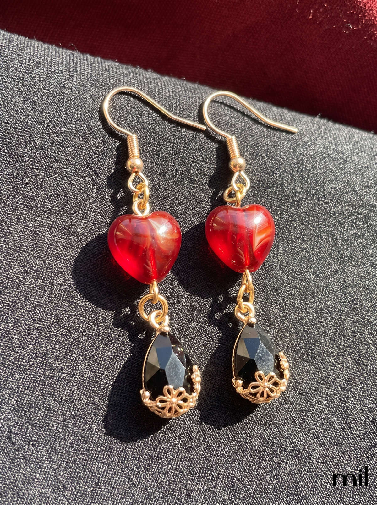 A pair of gold-tone dangle earrings with translucent red heart-shaped beads and faceted black teardrop charms, displayed on a textured dark gray surface.
