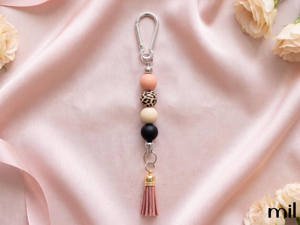 A beaded keychain with a silver carabiner clip, featuring pastel peach, leopard-print, cream, and black beads, a pink tassel, and a small gold accent, displayed on a draped pink satin fabric with decorative roses.