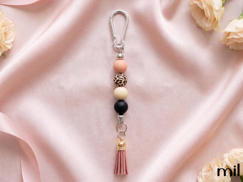 A beaded keychain with a silver carabiner clip, featuring pastel peach, leopard-print, cream, and black beads, a pink tassel, and a small gold accent, displayed on a draped pink satin fabric with decorative roses.