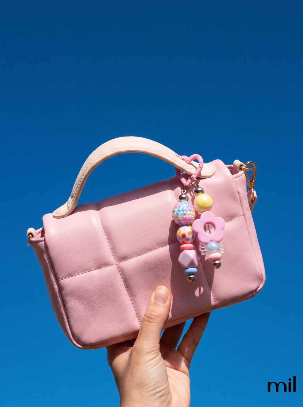 A hand holds a small pink quilted handbag against a clear blue sky. The bag has a light pink top handle, gold-tone hardware, and colorful beaded keychain charms hanging from the side.