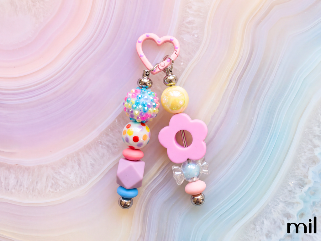 A pastel pink beaded keychain or bag charm with a heart-shaped top, dangling beads, and a flower detail, photographed on a soft pink and blue agate slab background. The brand name 'mil' is visible in the bottom right corner.