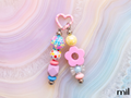 A pastel pink beaded keychain or bag charm with a heart-shaped top, dangling beads, and a flower detail, photographed on a soft pink and blue agate slab background. The brand name 'mil' is visible in the bottom right corner.