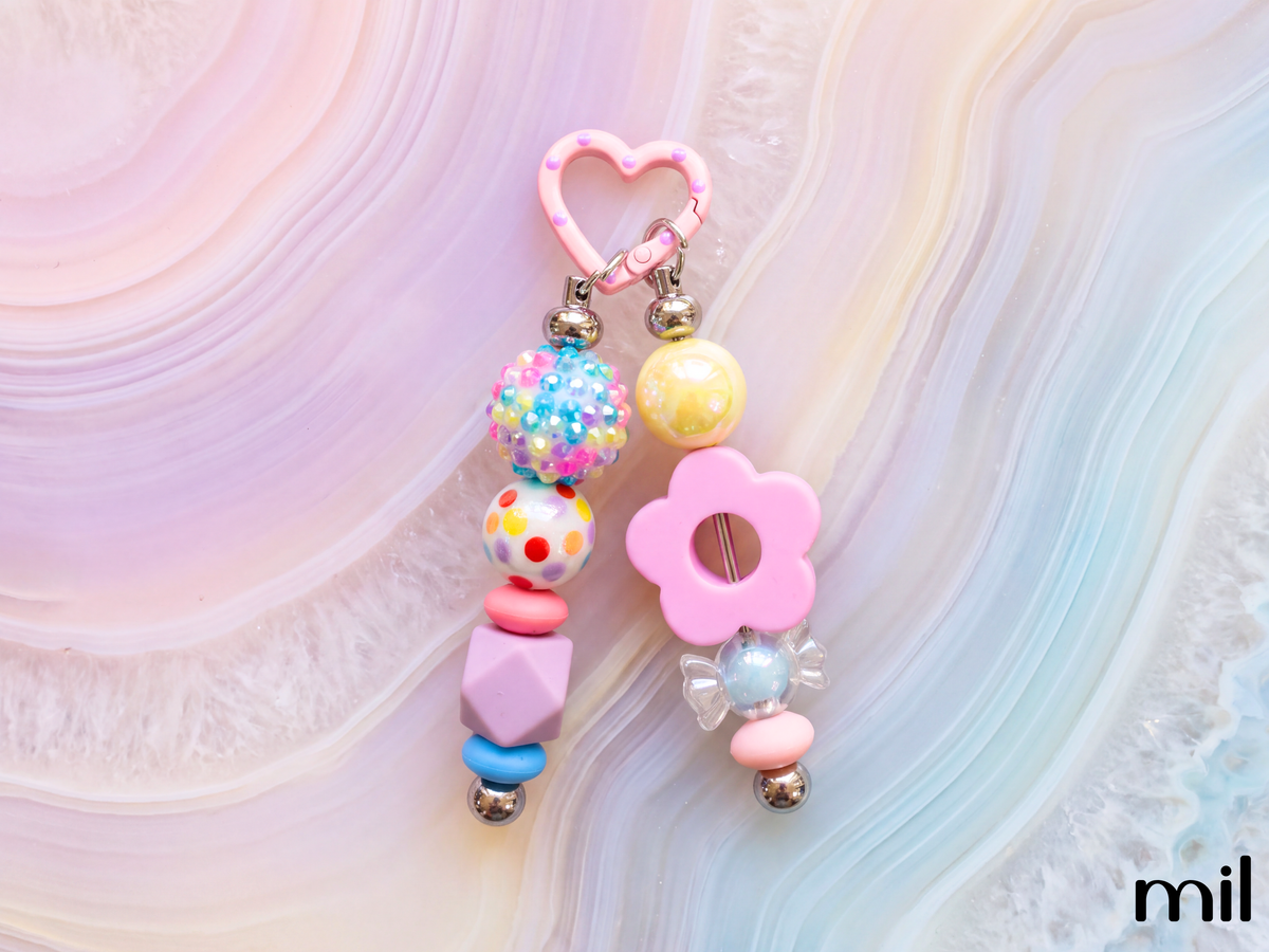 A pastel pink beaded keychain or bag charm with a heart-shaped top, dangling beads, and a flower detail, photographed on a soft pink and blue agate slab background. The brand name 'mil' is visible in the bottom right corner.