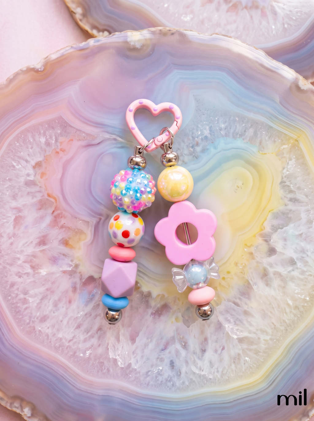 A pair of pastel beaded keychain bars displayed on a colorful agate slice. The earrings feature a pink heart-shaped clasp, a multicolor speckled bead, a yellow pearl-like bead, a pink flower charm, and pastel geometric beads in pink, blue, and purple.