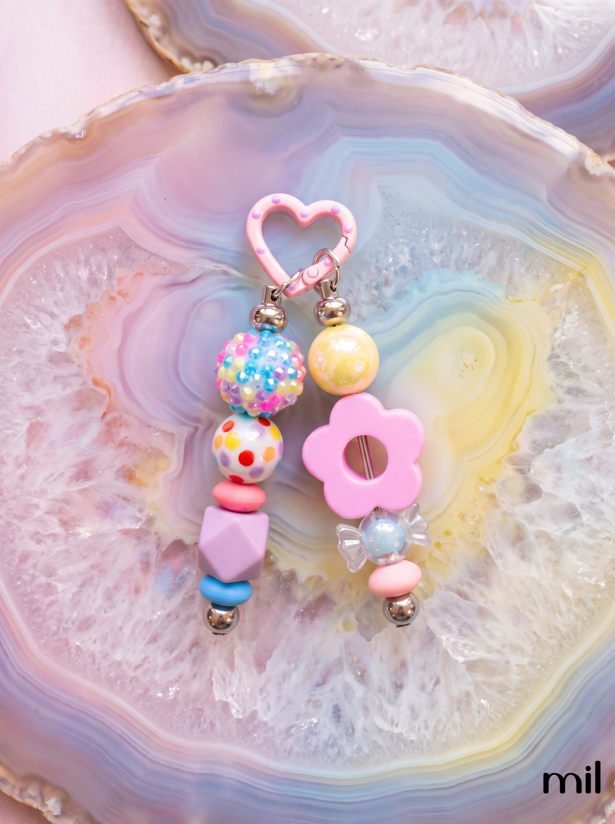 A pair of pastel beaded keychain bars displayed on a colorful agate slice. The earrings feature a pink heart-shaped clasp, a multicolor speckled bead, a yellow pearl-like bead, a pink flower charm, and pastel geometric beads in pink, blue, and purple.