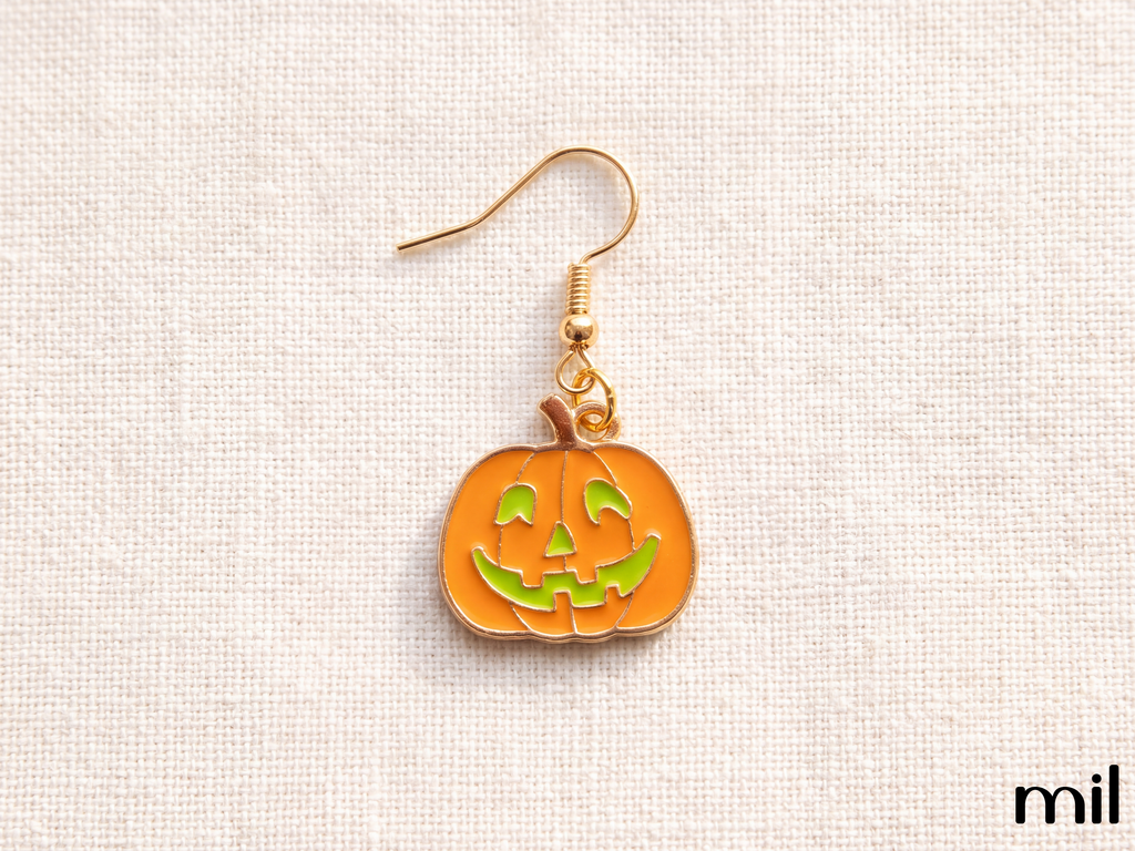 A single pumpkin-shaped dangling earring with an orange enamel body, carved jack-o'-lantern face in green, and a gold-tone fishhook ear wire, shown on a light beige textured fabric background.
