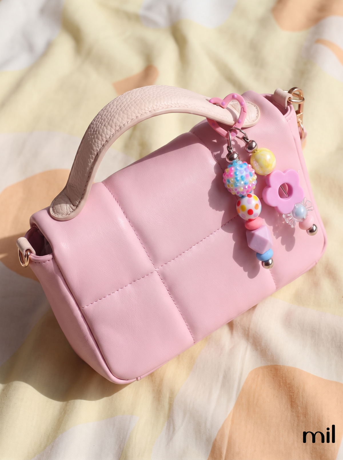 A pink quilted top-handle handbag with a colorful beaded charm keychain, resting on a soft beige and cream patterned fabric background.