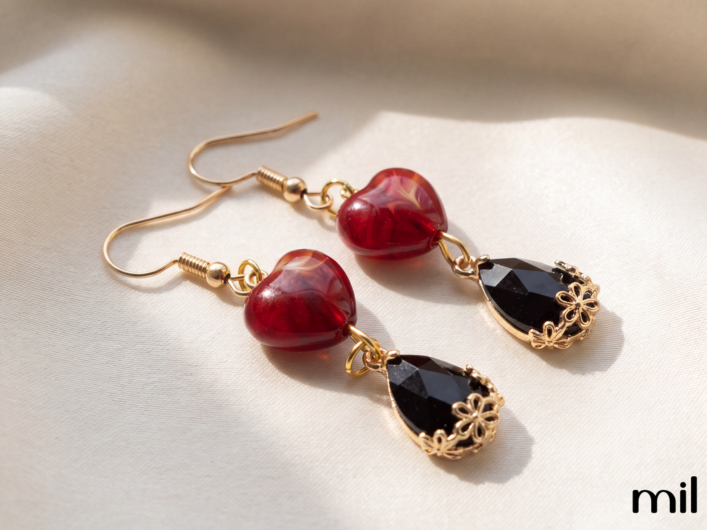 A pair of gold-tone dangle earrings with heart-shaped red beads and faceted black teardrop drops, displayed on a soft beige fabric background.