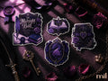 A styled flat-lay of four gothic-themed stickers or decals with purple and black designs, including a book, an open book, a journal, and an ornate crest with roses and a snake, arranged on a dark velvet surface with roses, a key, and ribbon.