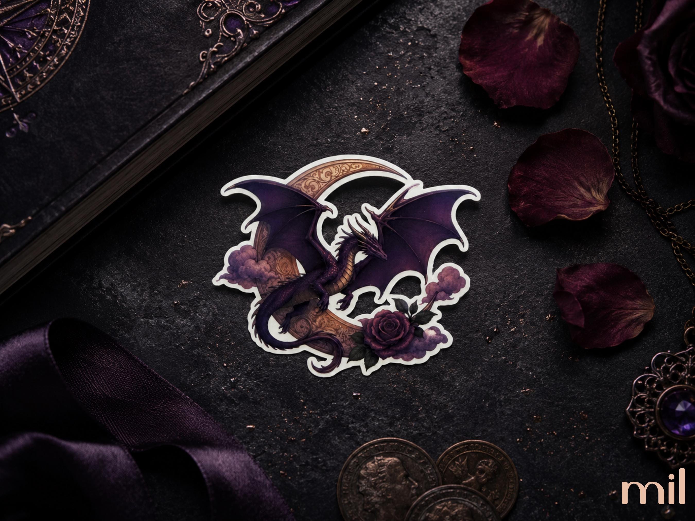 A die-cut sticker featuring a dark purple dragon wrapped around a crescent moon and rose, displayed on a dark textured surface with gothic props including dried rose petals, coins, and a dark book.