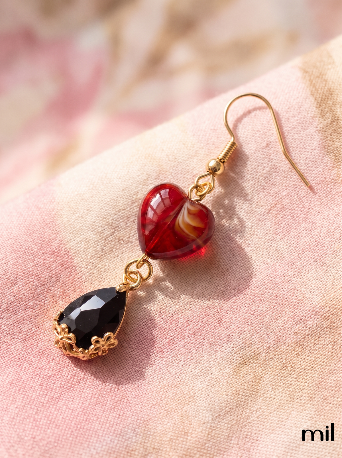 A pair of gold-tone dangle earrings displayed on a soft pink textured fabric. One earring features a polished red heart-shaped bead with a swirl of gold and white, and the other features a faceted black teardrop gemstone set in ornate gold filigree.