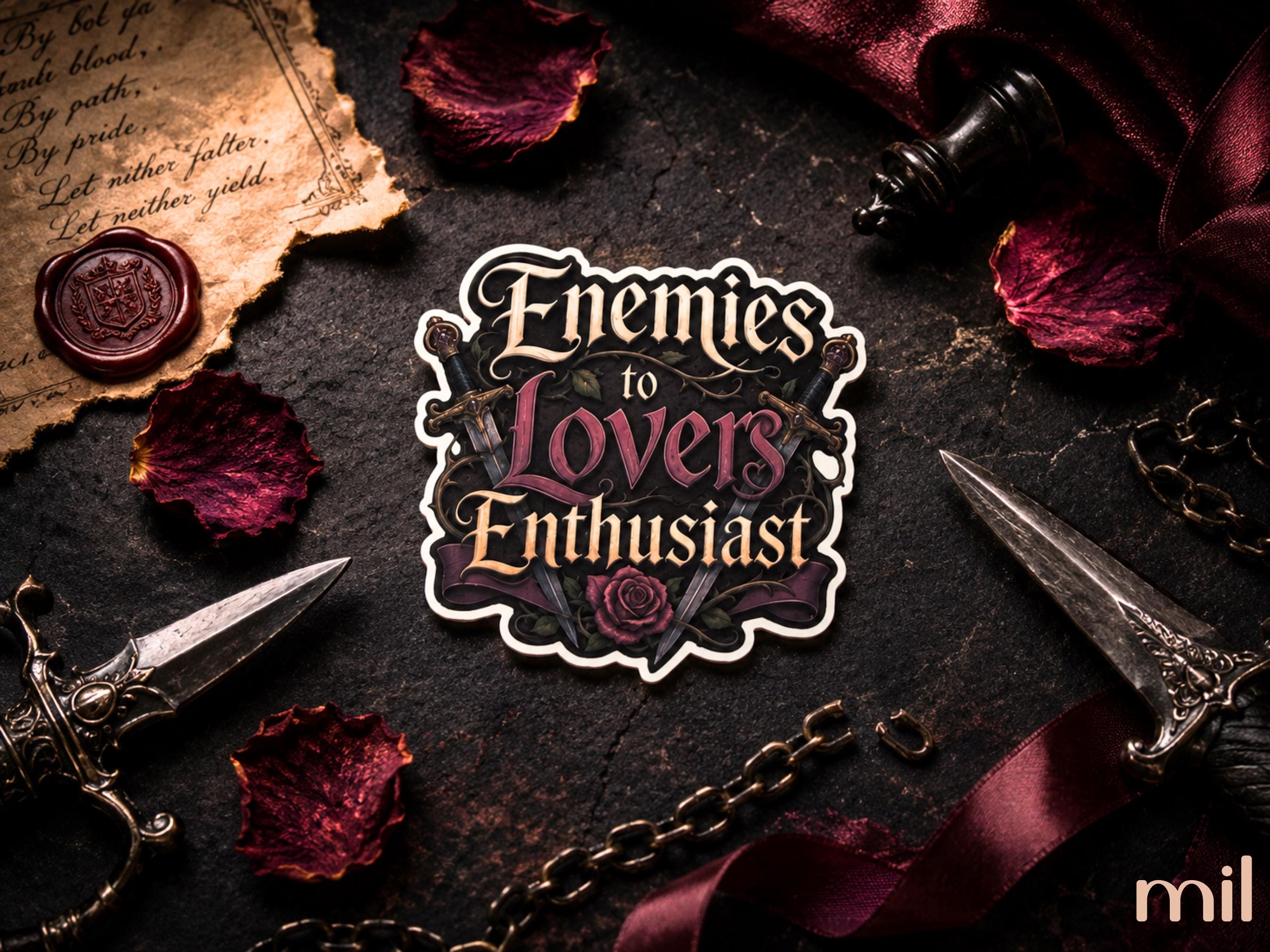 A die-cut sticker with the gothic text 'Enemies to Lovers Enthusiast' and a rose illustration, displayed on a dark textured surface surrounded by vintage props including a dagger, old paper, wax seal, chess piece, and red rose petals.