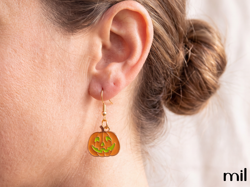 A close-up of a person's ear wearing a gold dangle earring with a carved orange pumpkin charm featuring green details, against a soft white background.