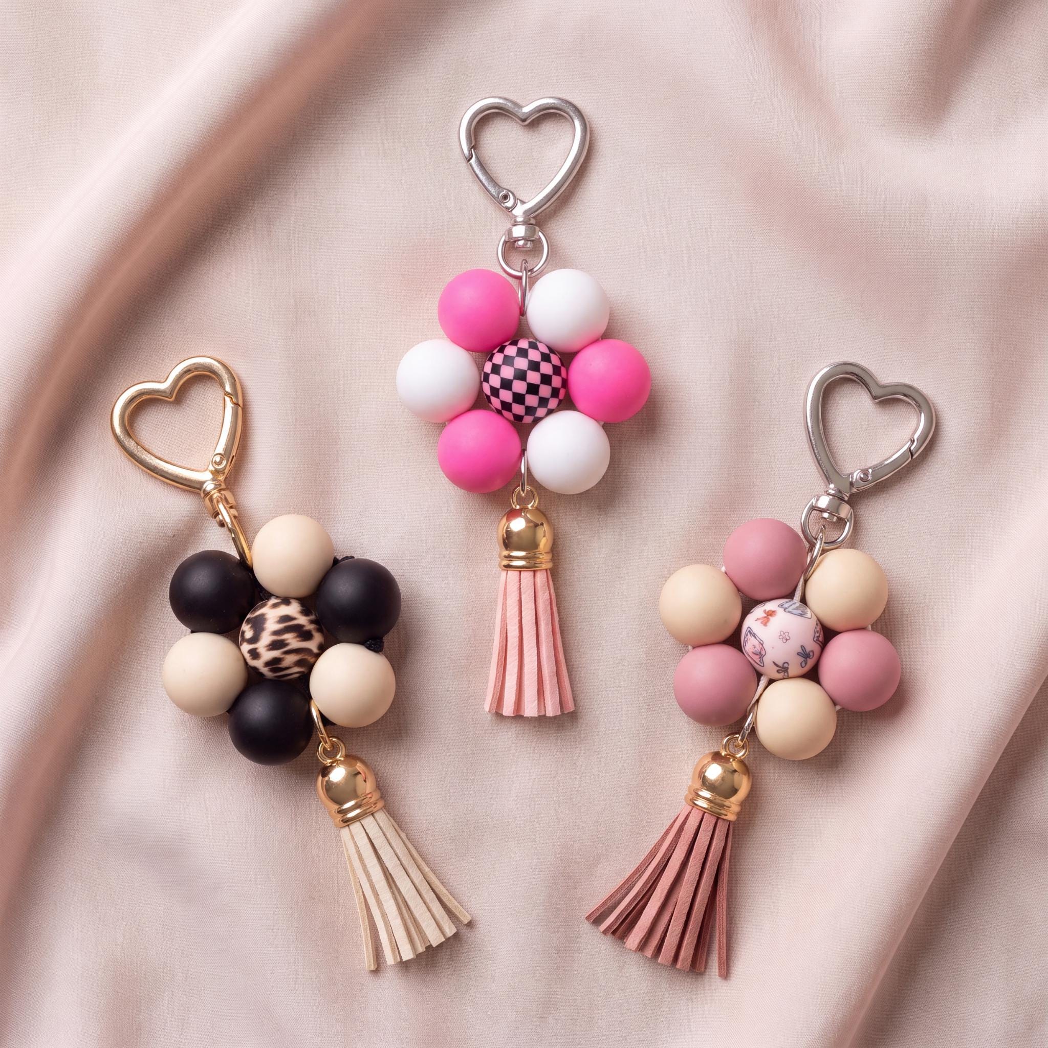 Three beaded keychains with heart-shaped metal rings and tassels, arranged on a soft pink satin fabric background. The keychains feature combinations of pink, white, black, beige, and leopard-print beads.