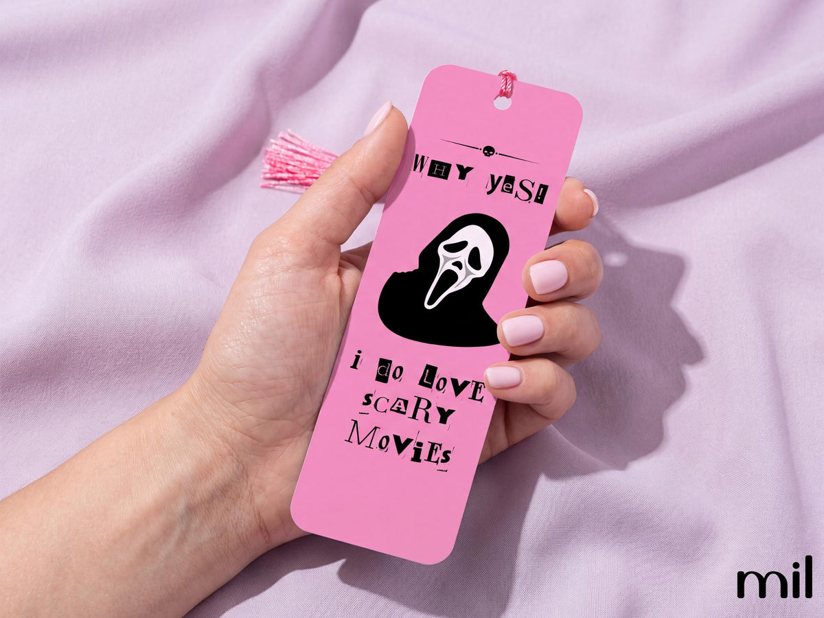 A hand holds a pink bookmark featuring a black Ghostface silhouette and the text 'WHY YES I DO LOVE SCARY MOVIES' against a soft lavender fabric background.