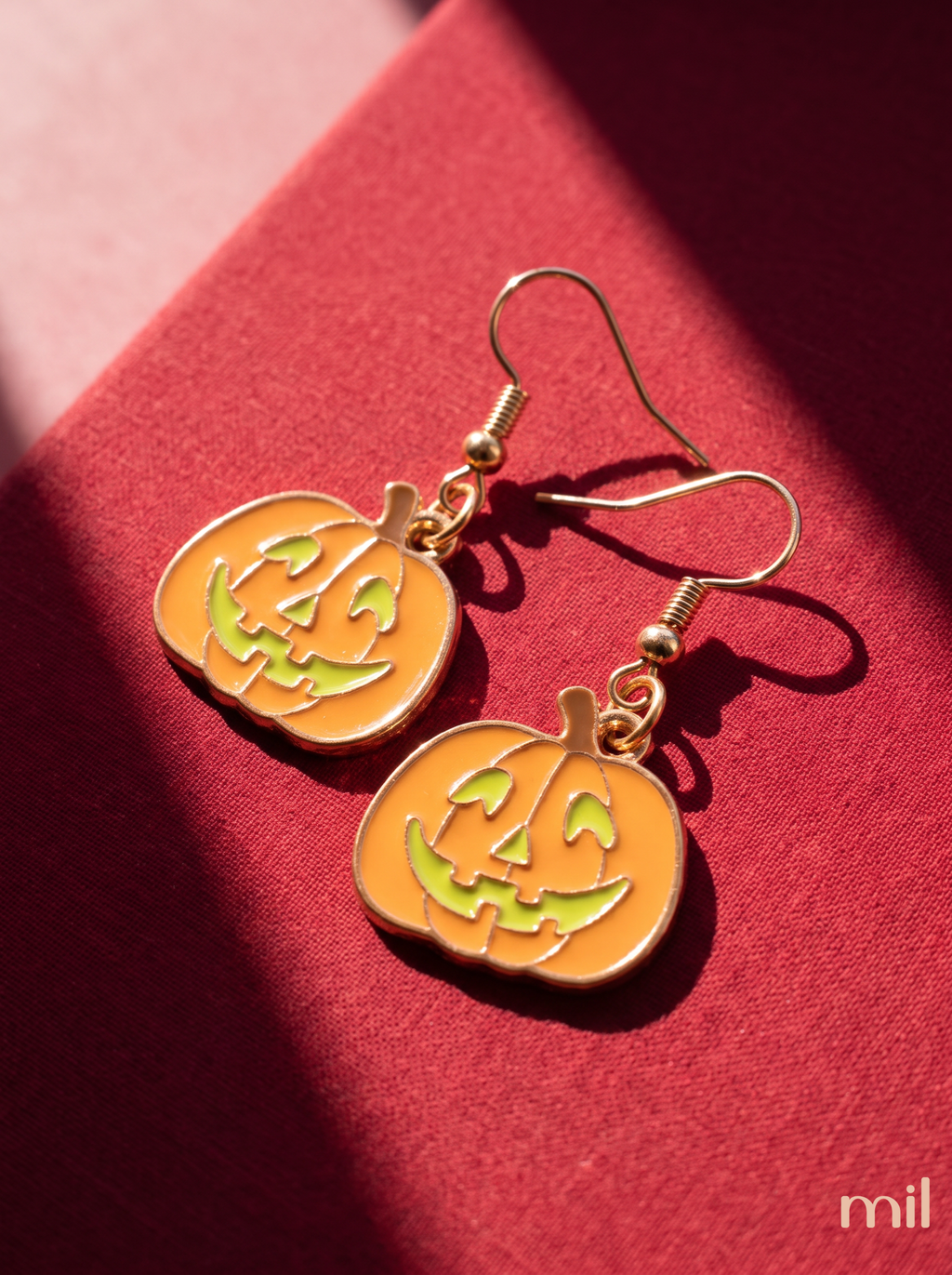 Gold-tone pumpkin-shaped dangle earrings with orange enamel and green jack-o'-lantern facial details, displayed on a textured red surface with dramatic shadows.