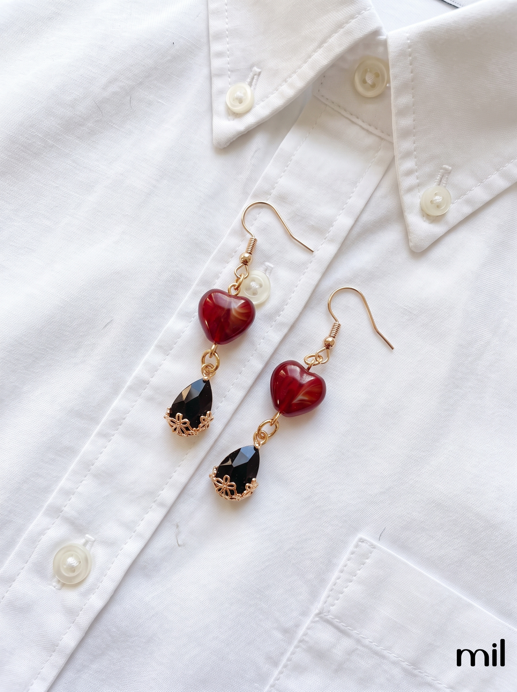 A pair of gold-tone dangle earrings with red heart-shaped beads and black teardrop charms, displayed on a white button-up shirt.